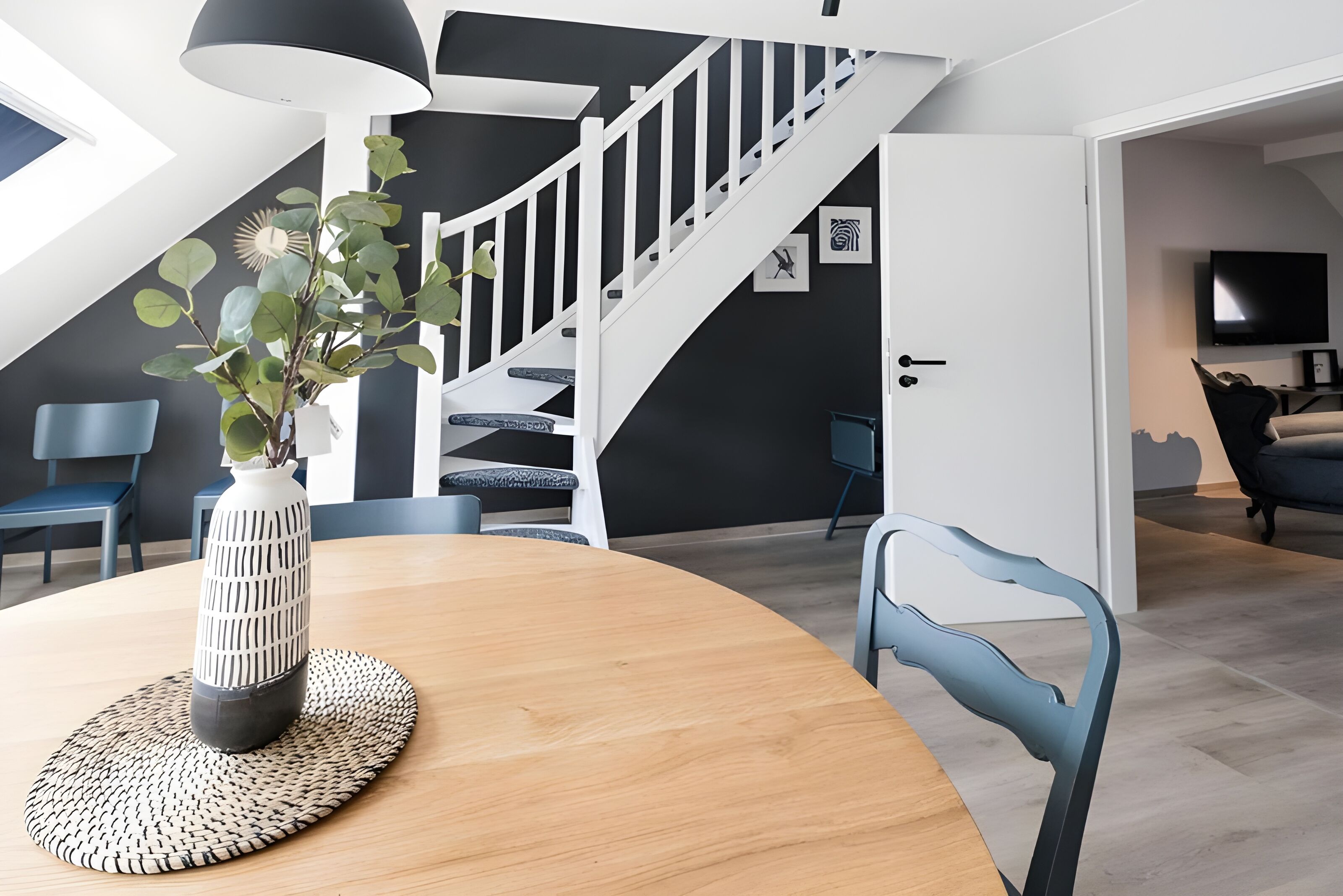 Un salon moderne avec un escalier, une table à manger agrémentée d’un vase et des chaises bleues.