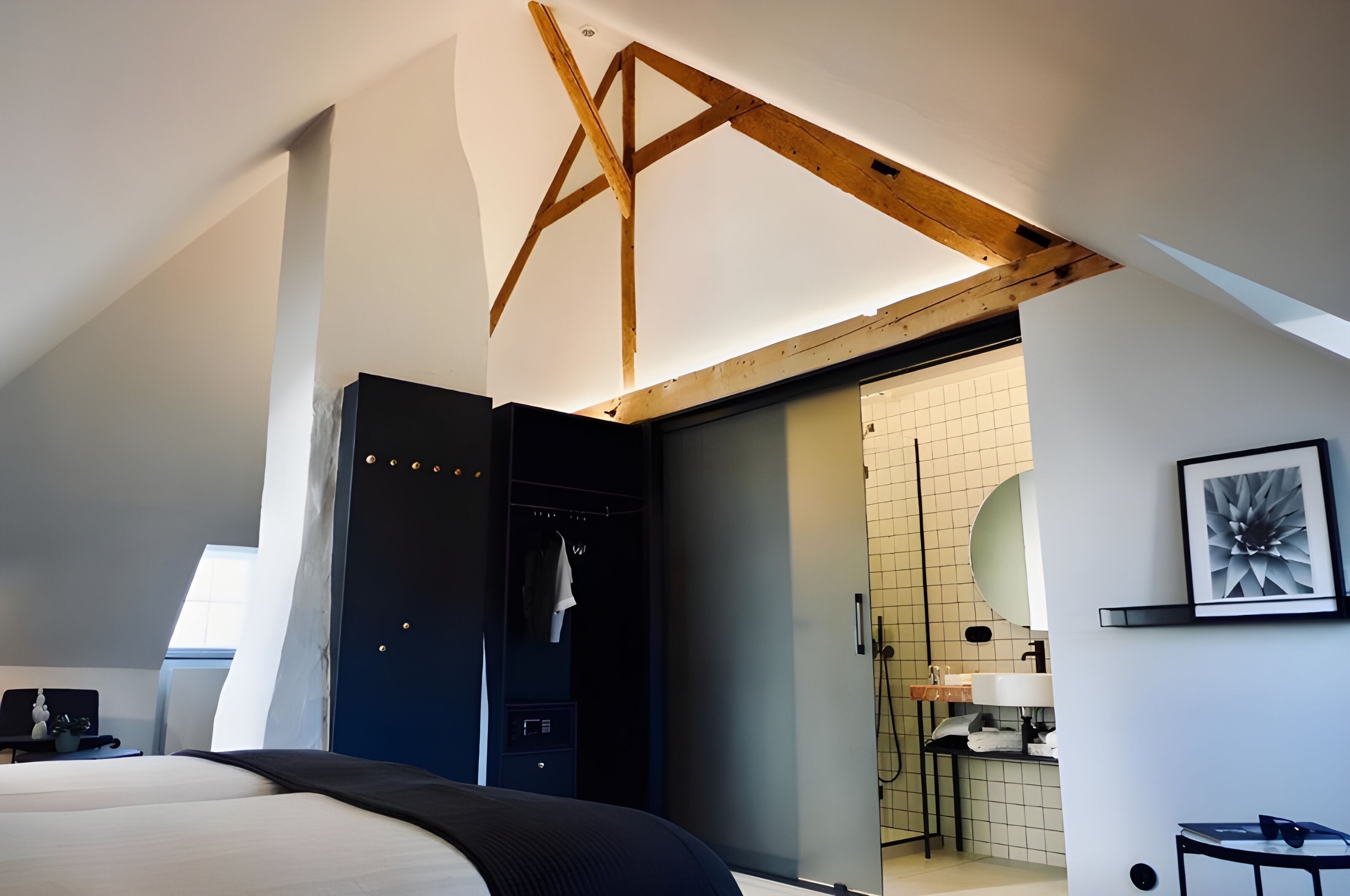 A modern hotel room with a sloped ceiling, a walk-in closet, a bathroom with a tiled wall, and a framed picture on the wall.
