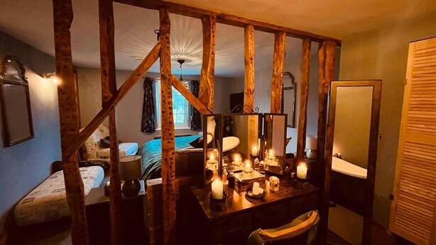Um quarto aconchegante com vigas de madeira aparentes, uma cama, uma escrivaninha com velas e um espelho.