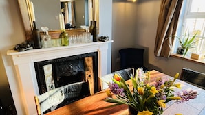 Uma área de refeições com uma mesa de madeira, um vaso de flores, uma lareira com um espelho acima e uma janela com cortinas.
