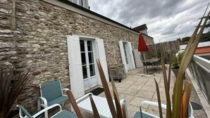 A stone building with a sloped roof, a small window, and a door. There are outdoor chairs and a table, along with a red umbrella.