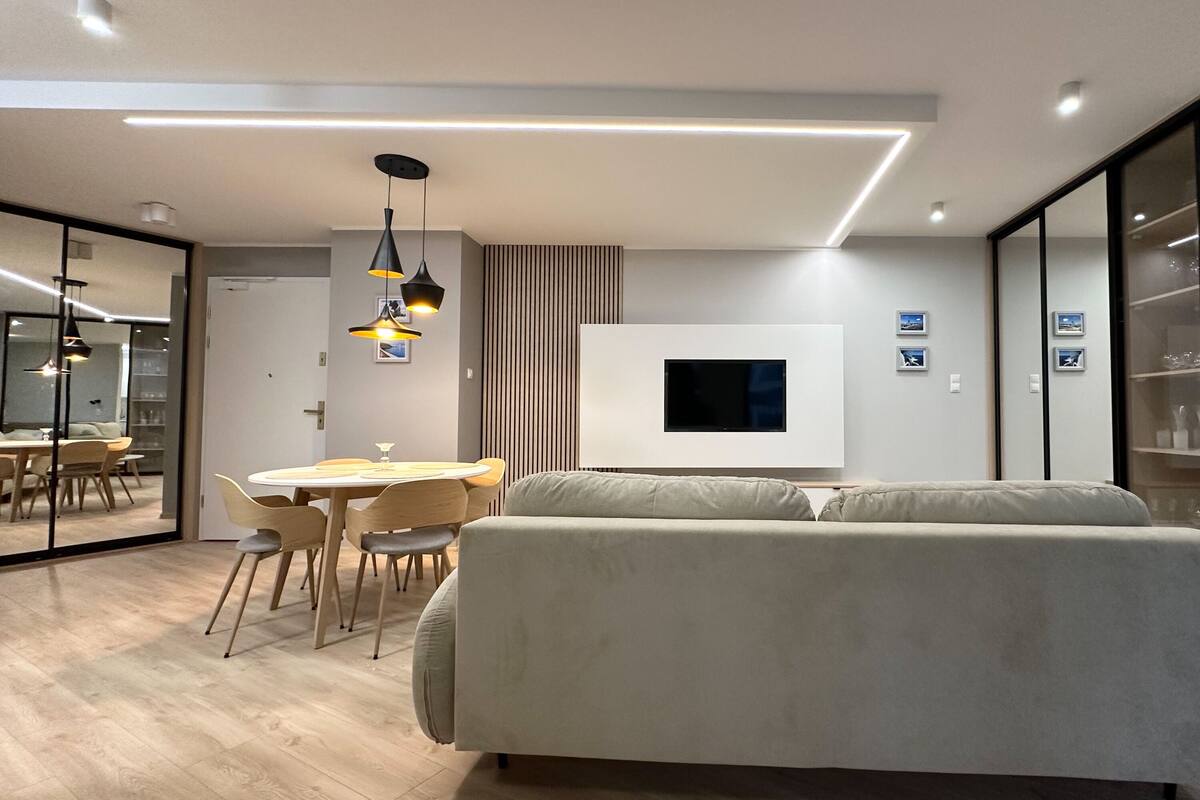 A modern living room with a sofa, dining table, and chairs. There is a flat-screen TV mounted on the wall and pendant lights above the table.