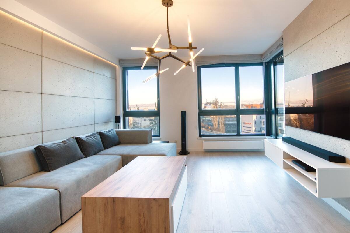 A modern living room with a sectional sofa, a wooden coffee table, a large TV, and large windows offering a city view.