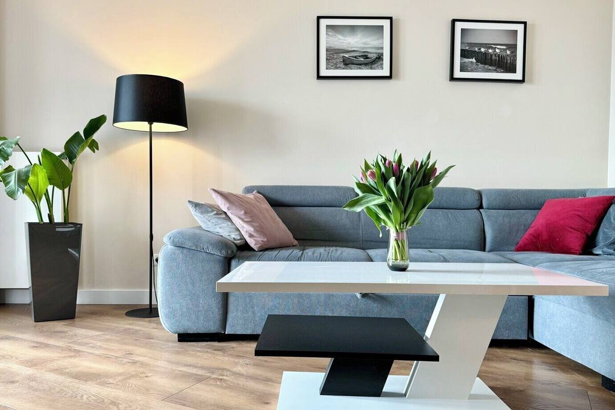 A modern living room with a grey sofa, a white coffee table, a black floor lamp, and two framed black and white photographs on the wall.