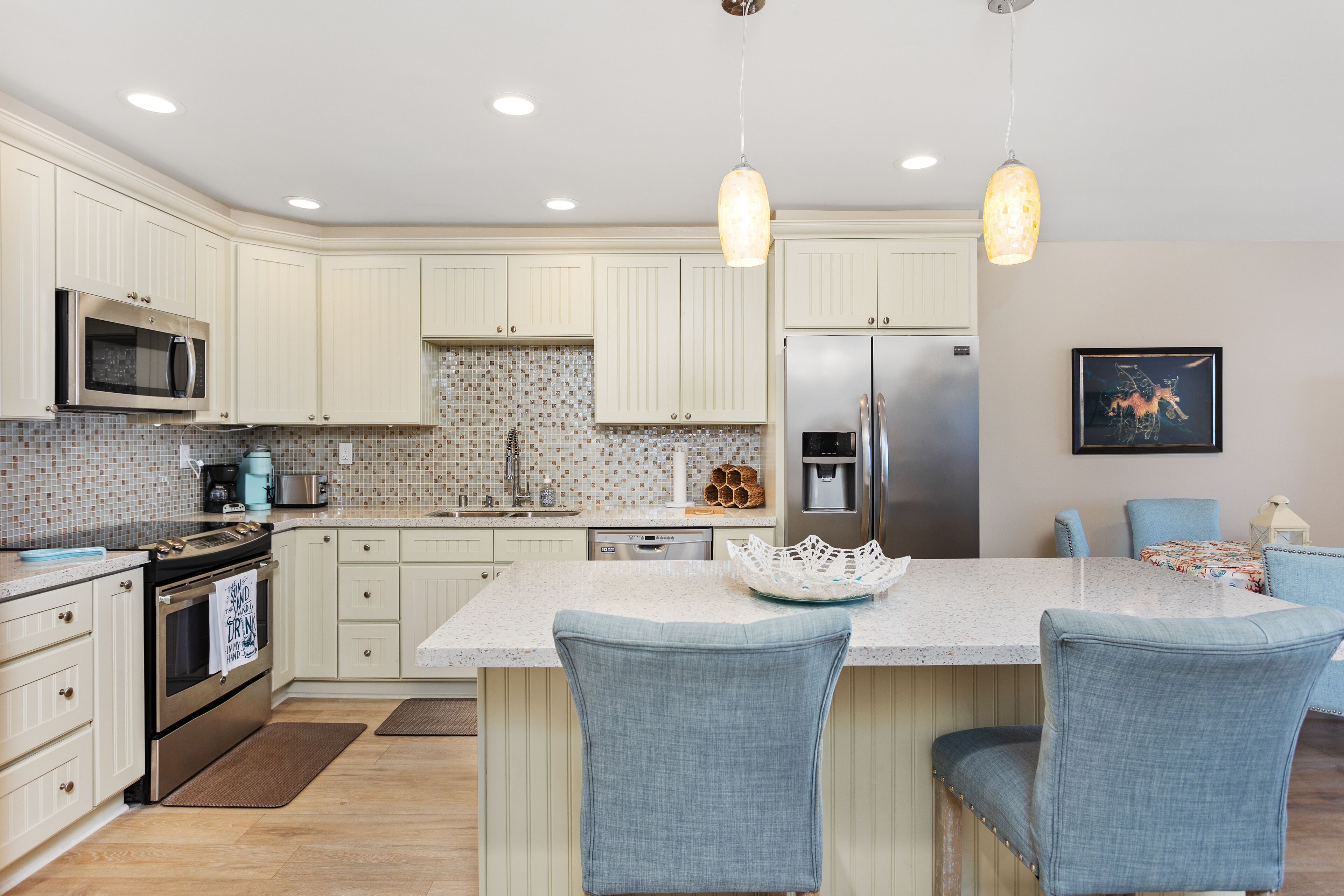 A kitchen with white cabinets, a stainless steel refrigerator, a microwave, and a sink. There are two blue chairs at the island and a hanging light fixture.