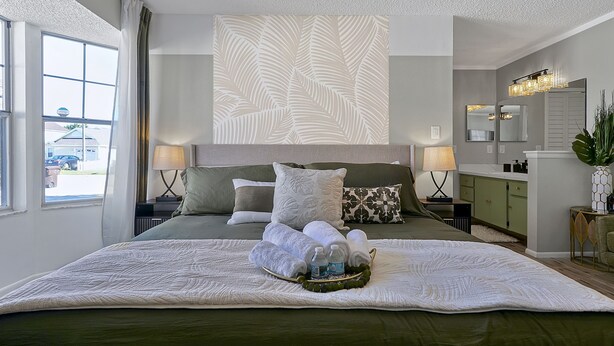 A neatly made bed with a green bedspread, a decorative leaf-patterned wall, and a bathroom with a mirror and lights. - Disney Villa w Priv Pool & Lake View-12 Mi parks Top 1% Listing on A i r b n b (Kissimmee)