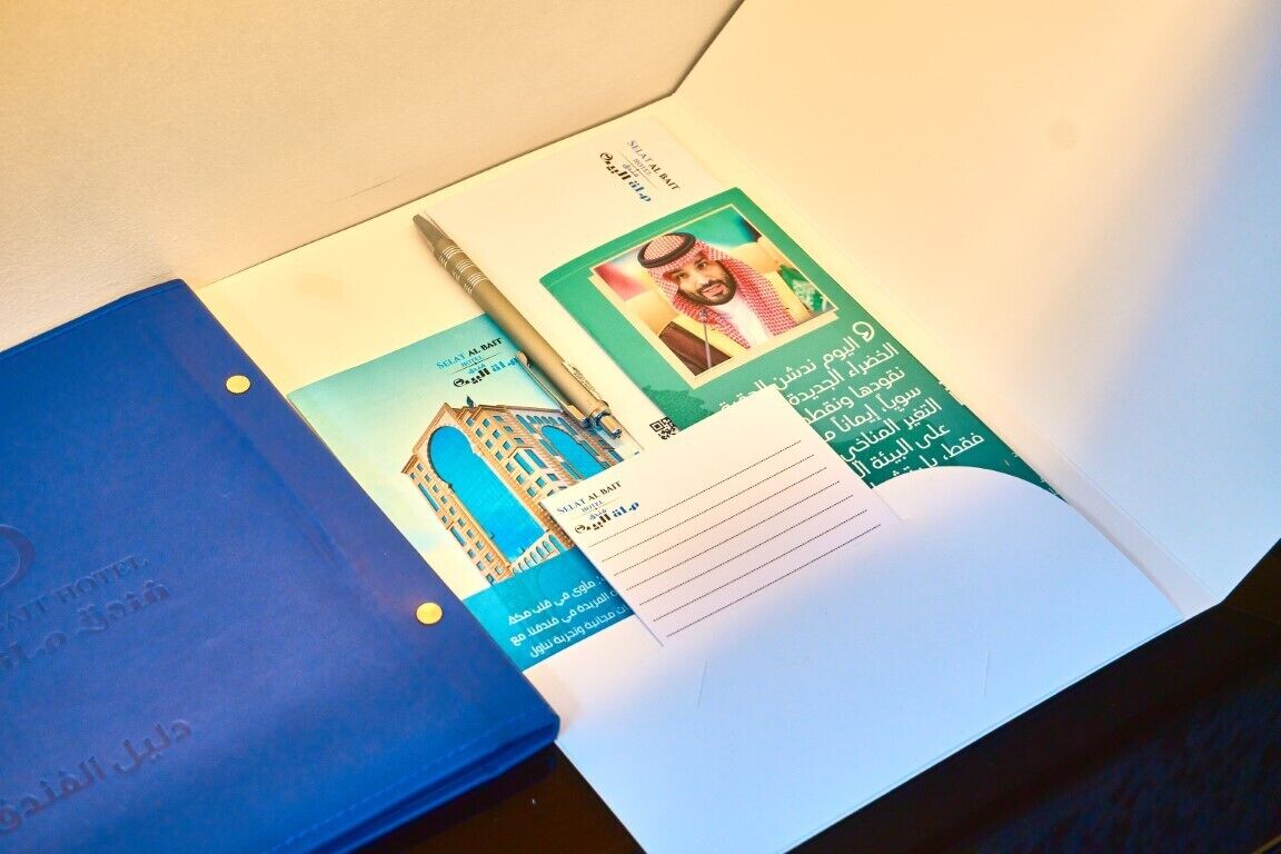 Un classeur bleu aux couleurs de l’hôtel, un stylo et une brochure contenant du texte en arabe et une photo d’un bâtiment.
