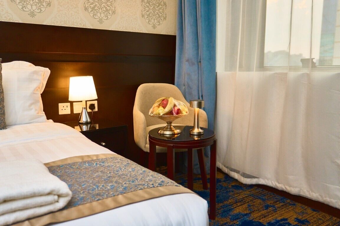 A hotel room with a neatly made bed, a small table with a lamp, a chair, and a view of the outside through a window with curtains.