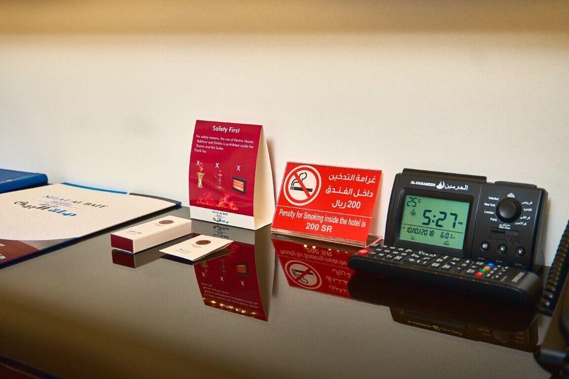 A hotel reception desk with a digital clock displaying 5:27, a telephone, and signs indicating safety and smoking policies.