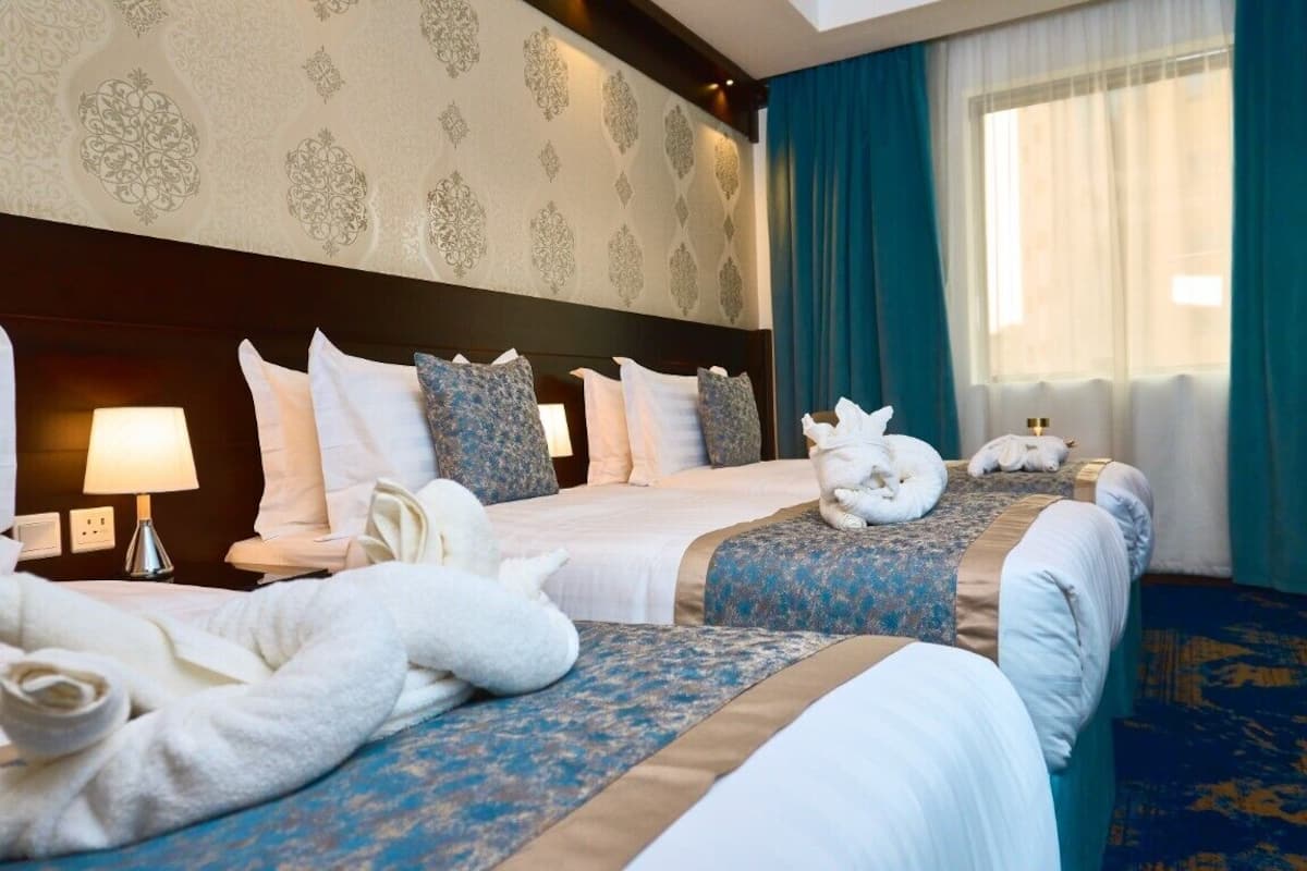 A hotel room with two beds, a patterned wallpaper, a large window with curtains, and a blue carpet.