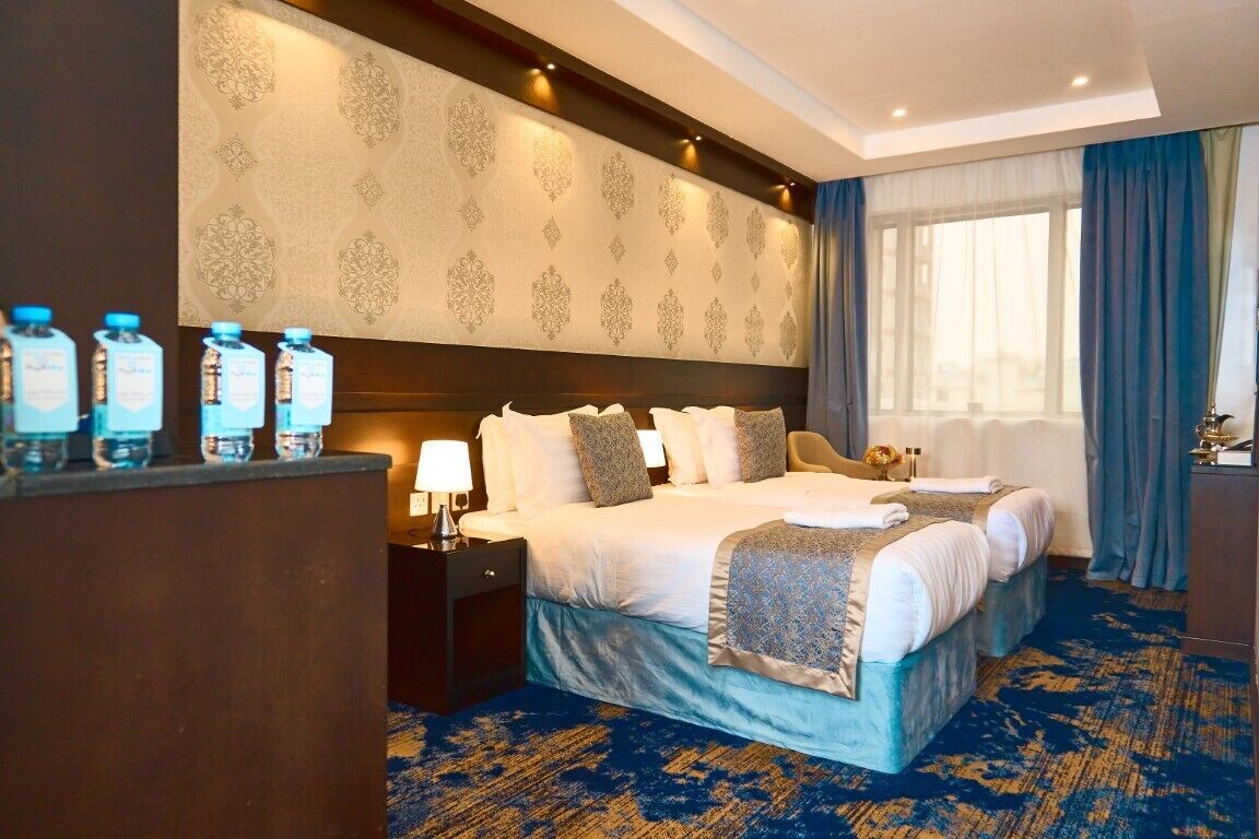 A hotel room with a large bed, two bedside tables, a nightstand, a window with curtains, and a blue carpet.