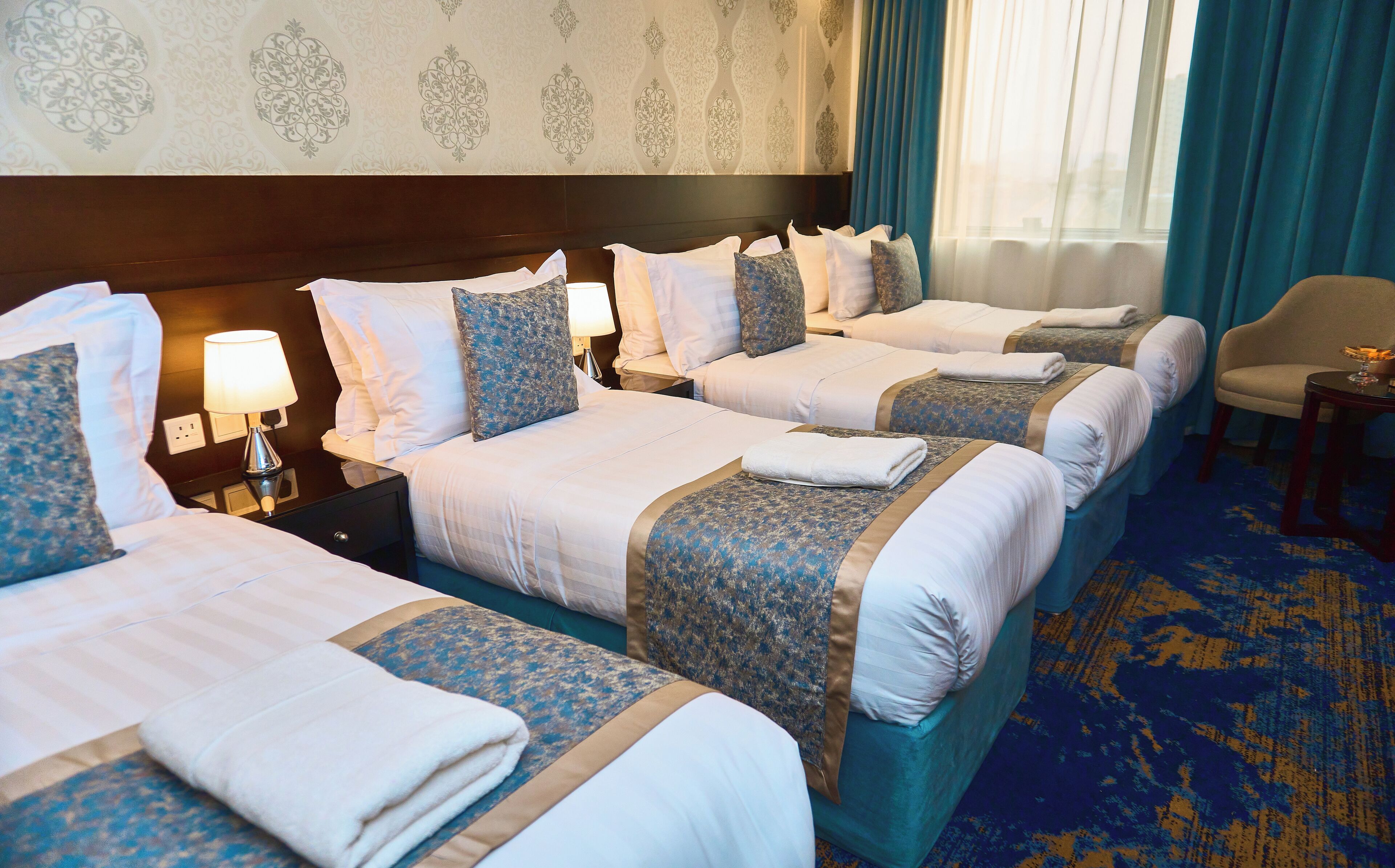 A hotel room with four beds, each with a blue and white bedspread, a headboard, and a nightstand with a lamp.