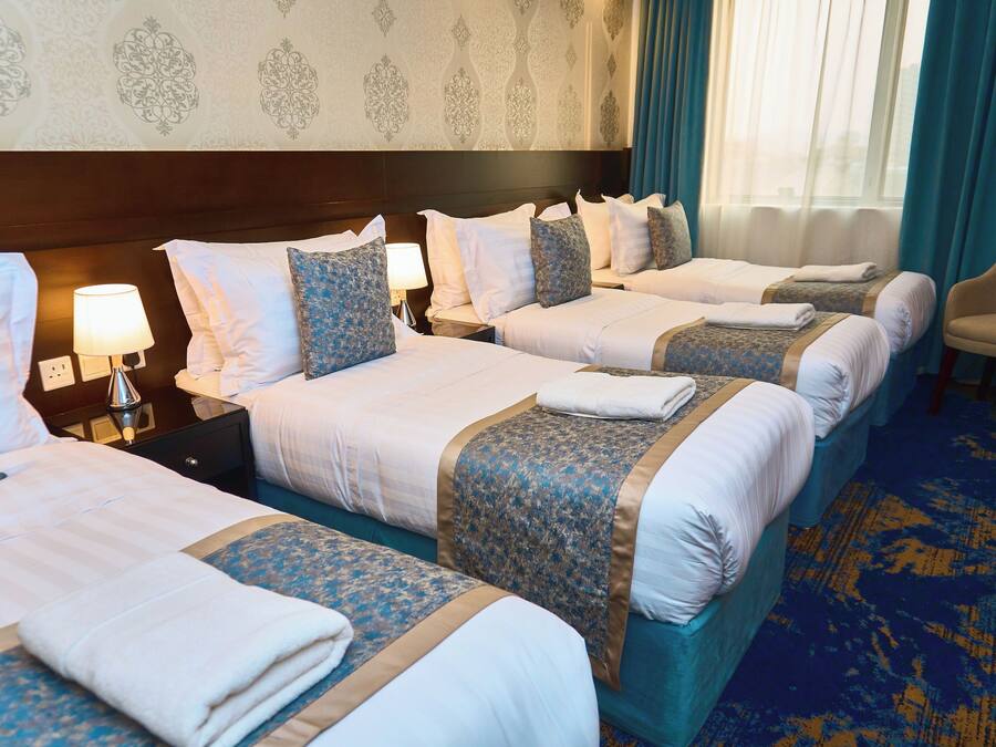 A hotel room with four beds, each with a blue and white bedspread, a headboard, and a nightstand with a lamp.