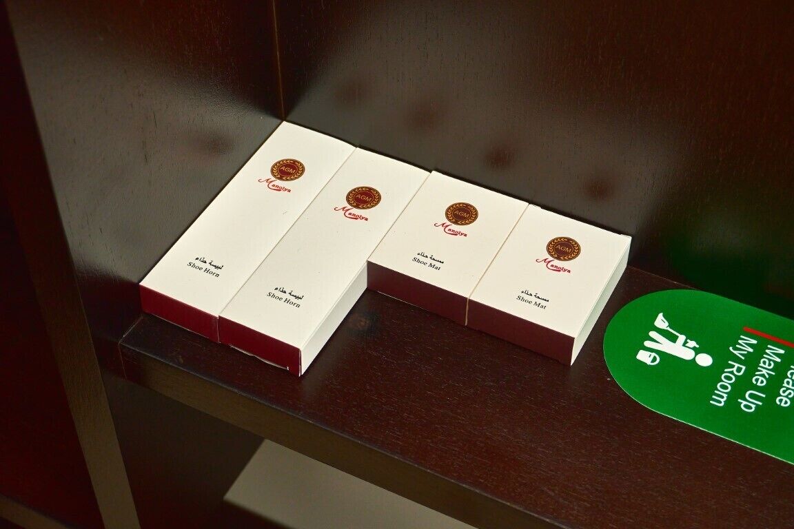 Four white boxes with red tops and gold logos, labeled "AIGI" and "Shoe Horn," on a wooden surface. A green sign with white text reads "Make Up Room."