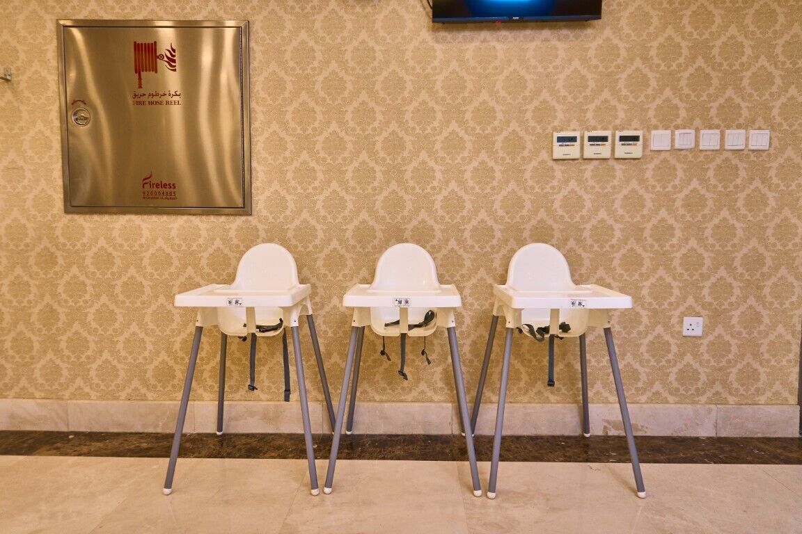Three white high chairs with metal legs against a patterned wall, a fire hose reel sign, and a television mounted above.