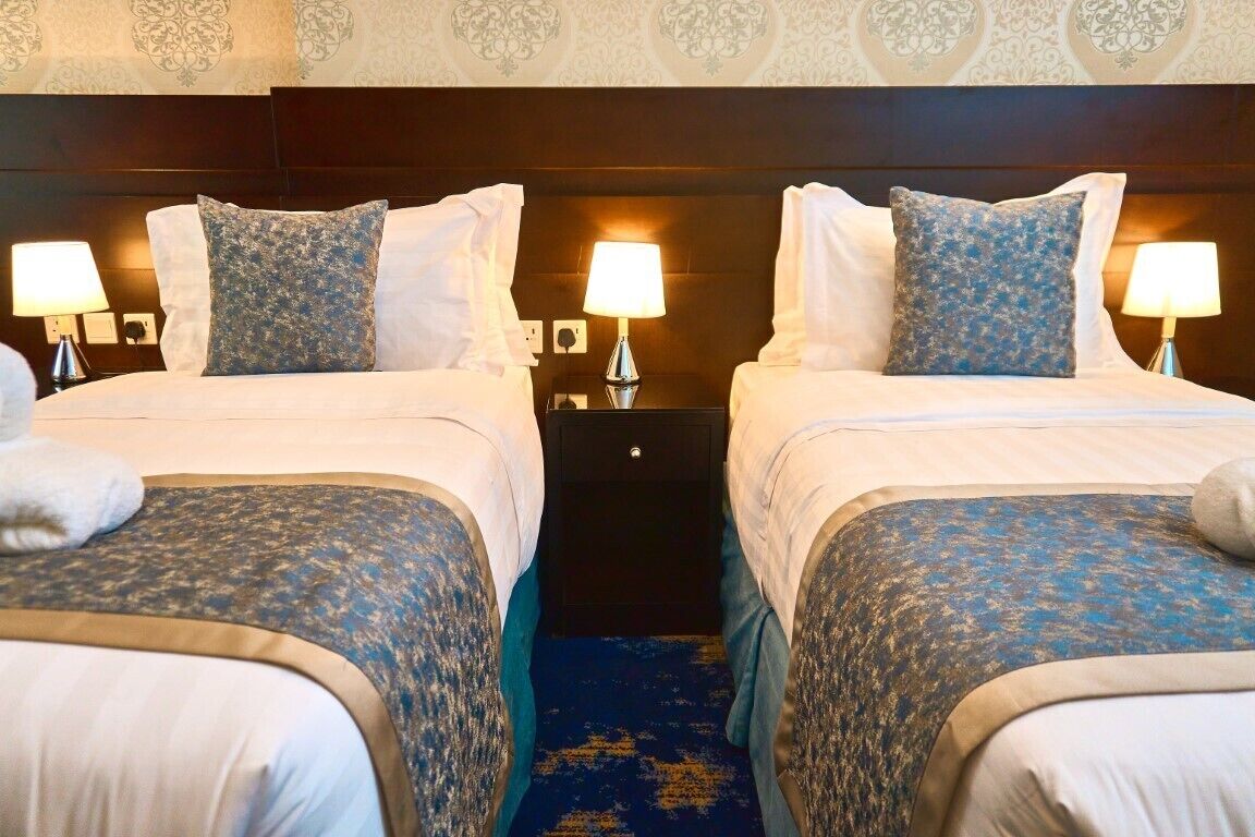 A hotel room with two beds, each with a patterned bedspread and pillows, a nightstand with a lamp, and a blue carpet.