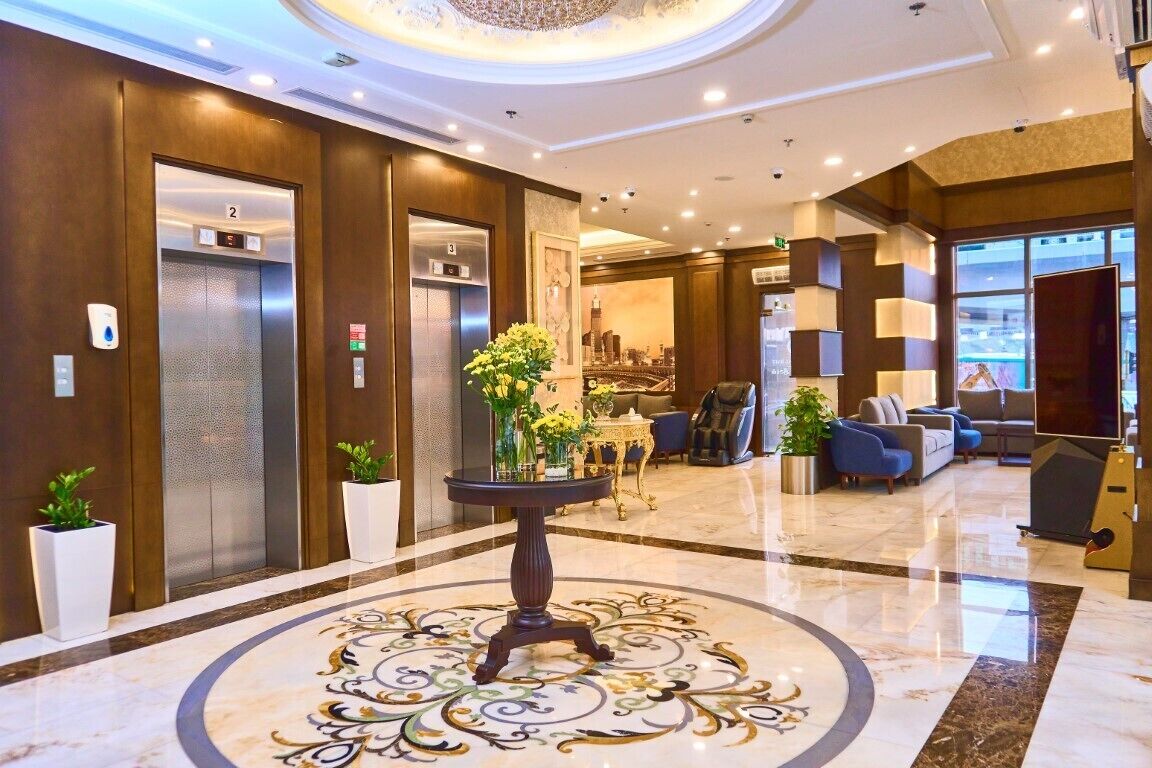 A hotel lobby with two elevators, a round marble table with a floral centerpiece, and a seating area with sofas and a television.