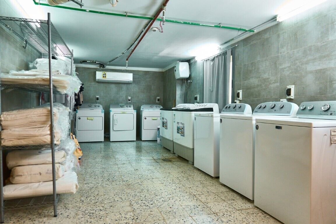 A laundry room with multiple washing machines and dryers, a metal shelf with folded linens, and an air conditioning unit on the wall.