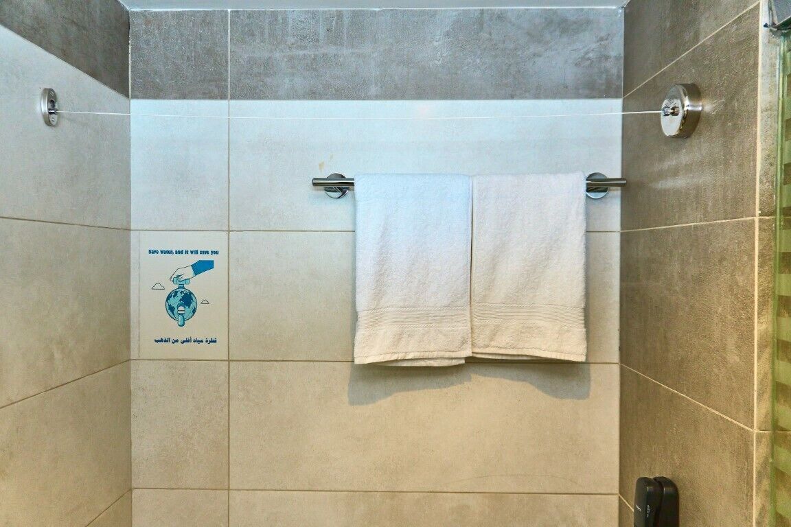 Zona de ducha con dos toallas blancas colgadas en una barra metálica, un mando visible para el control del agua y un cartel en la pared que promueve el ahorro de agua.