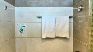 Zona de ducha con dos toallas blancas colgadas en una barra metálica, un mando visible para el control del agua y un cartel en la pared que promueve el ahorro de agua.