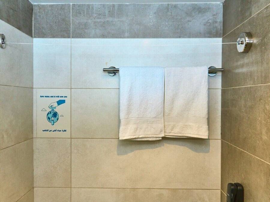 Zona de ducha con dos toallas blancas colgadas en una barra metálica, un mando visible para el control del agua y un cartel en la pared que promueve el ahorro de agua.