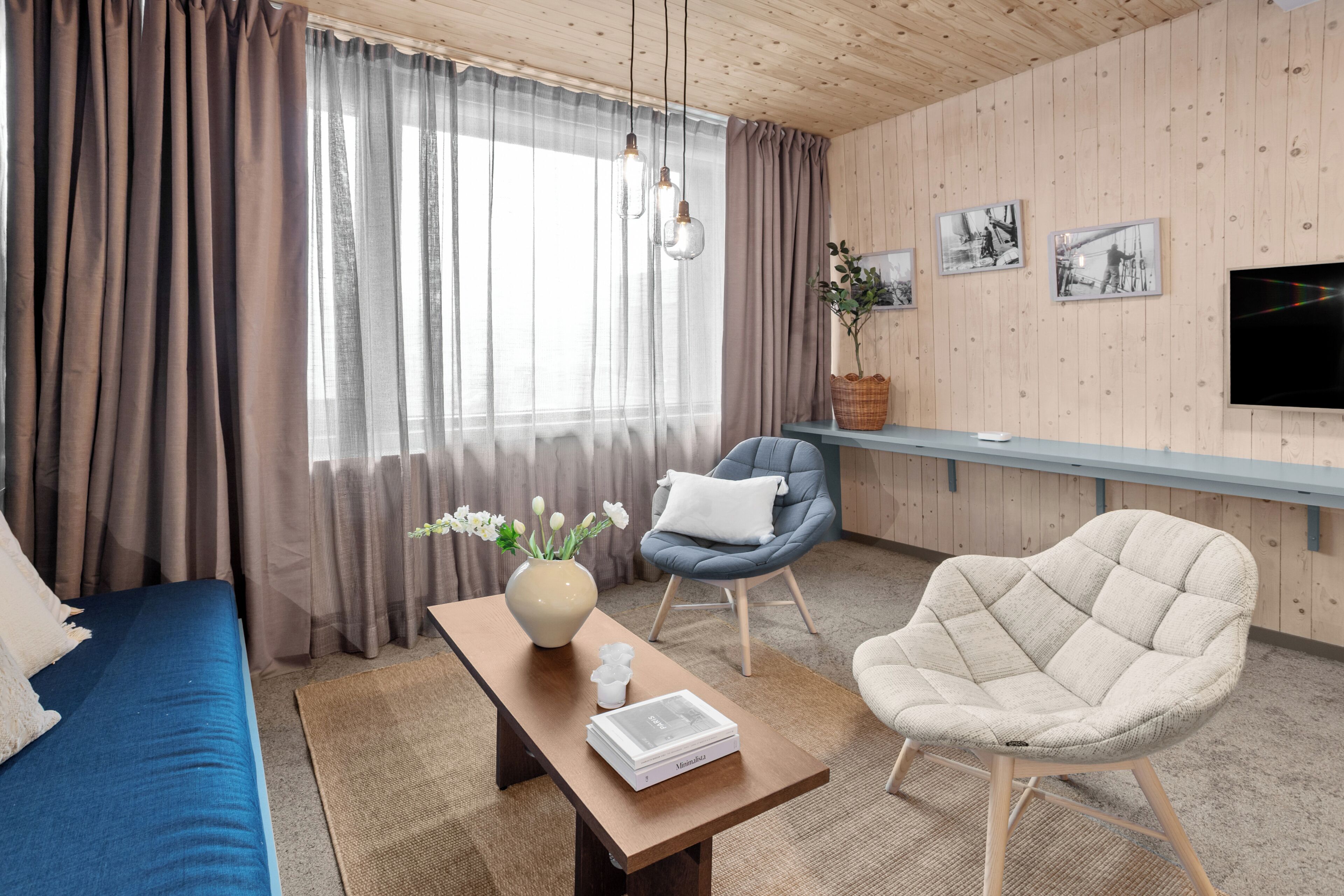 Una habitación con un sofá azul, una mesa de centro de madera con un libro, una silla azul, un sillón beige y un televisor montado en la pared.