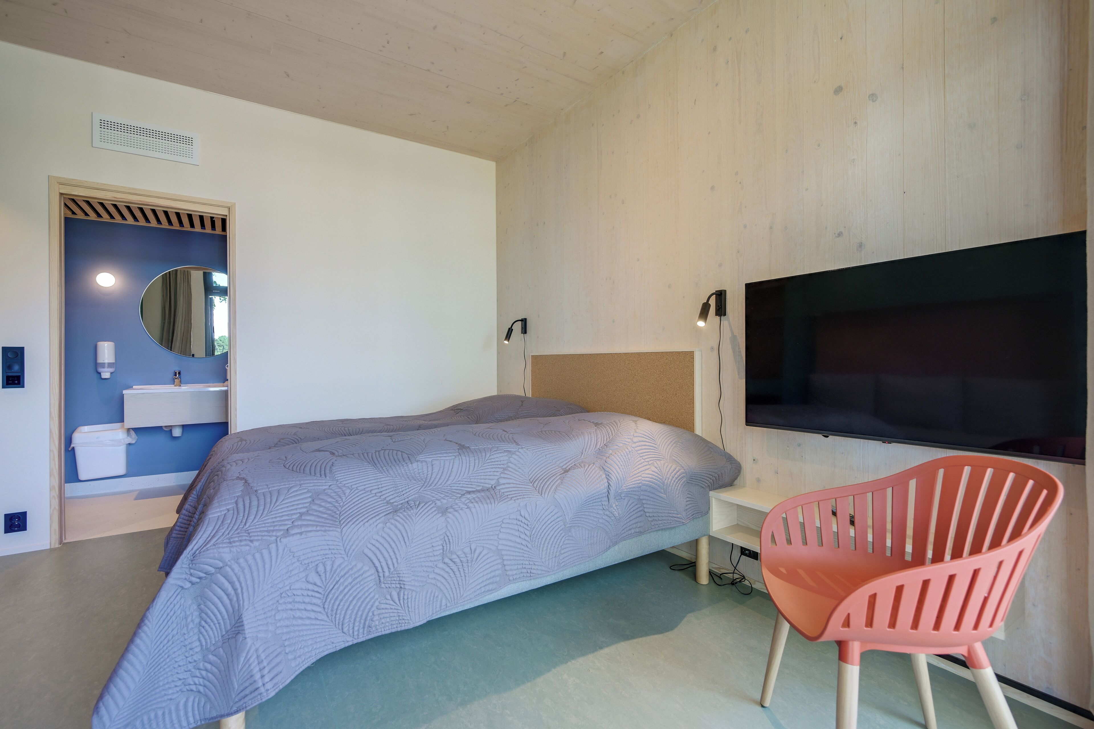 Une chambre à coucher avec un lit, une télévision, un fauteuil rose et une salle de bain visible à travers une porte ouverte.