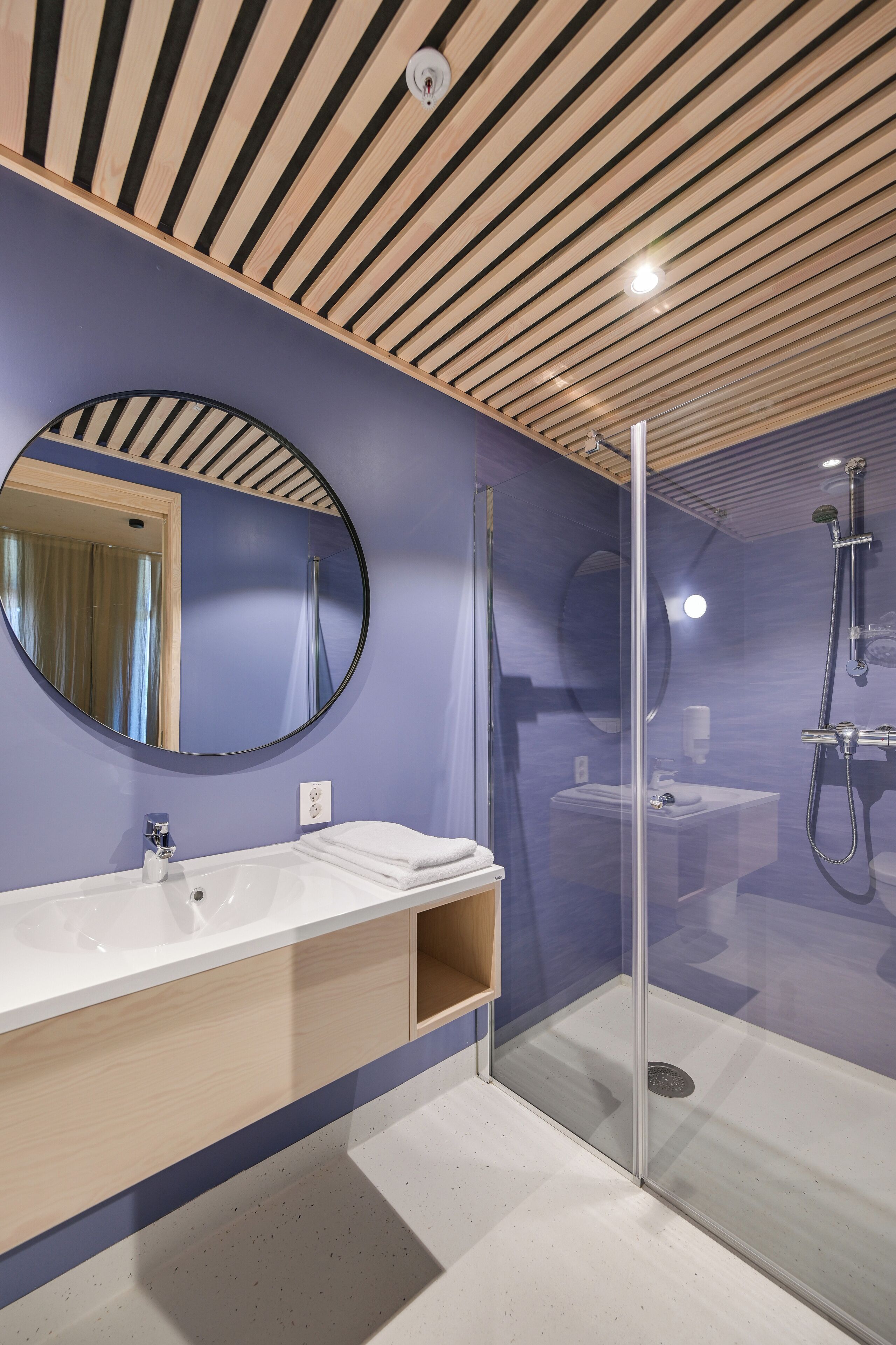 Un baño moderno con techo de madera, un espejo redondo grande, un lavabo blanco y una zona de ducha con mampara de vidrio.