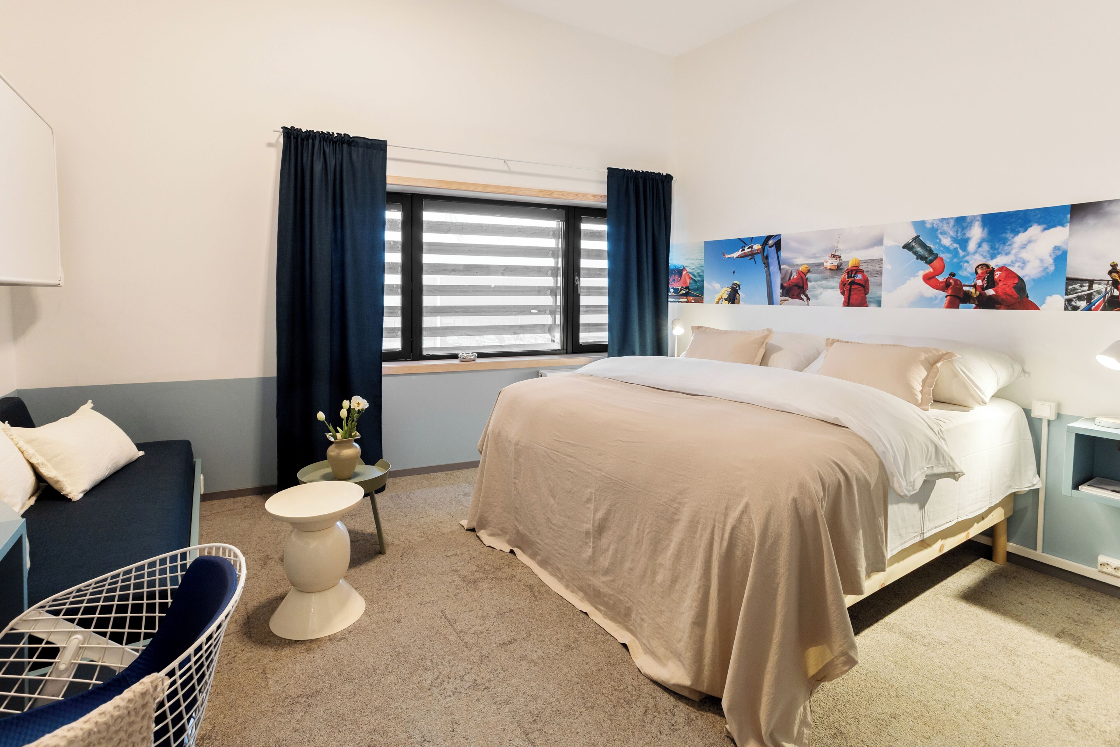 Quarto de hotel com cama, cadeira, mesa pequena, janela com cortinas e parede com várias fotografias.