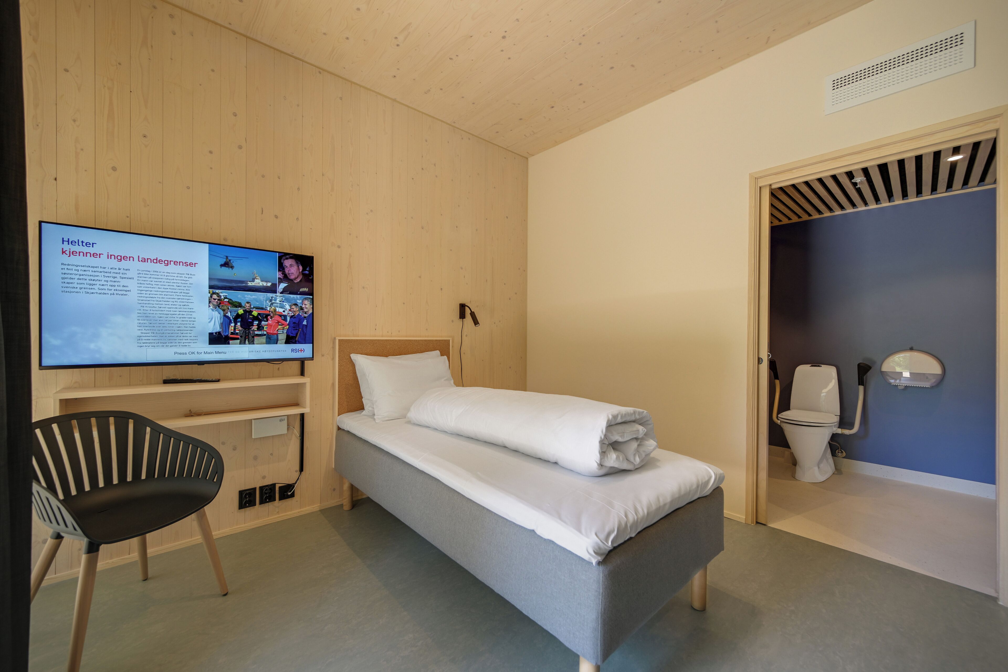 Una cama individual con colchón blanco, un televisor en la pared, una silla y un baño con inodoro y lavabo.