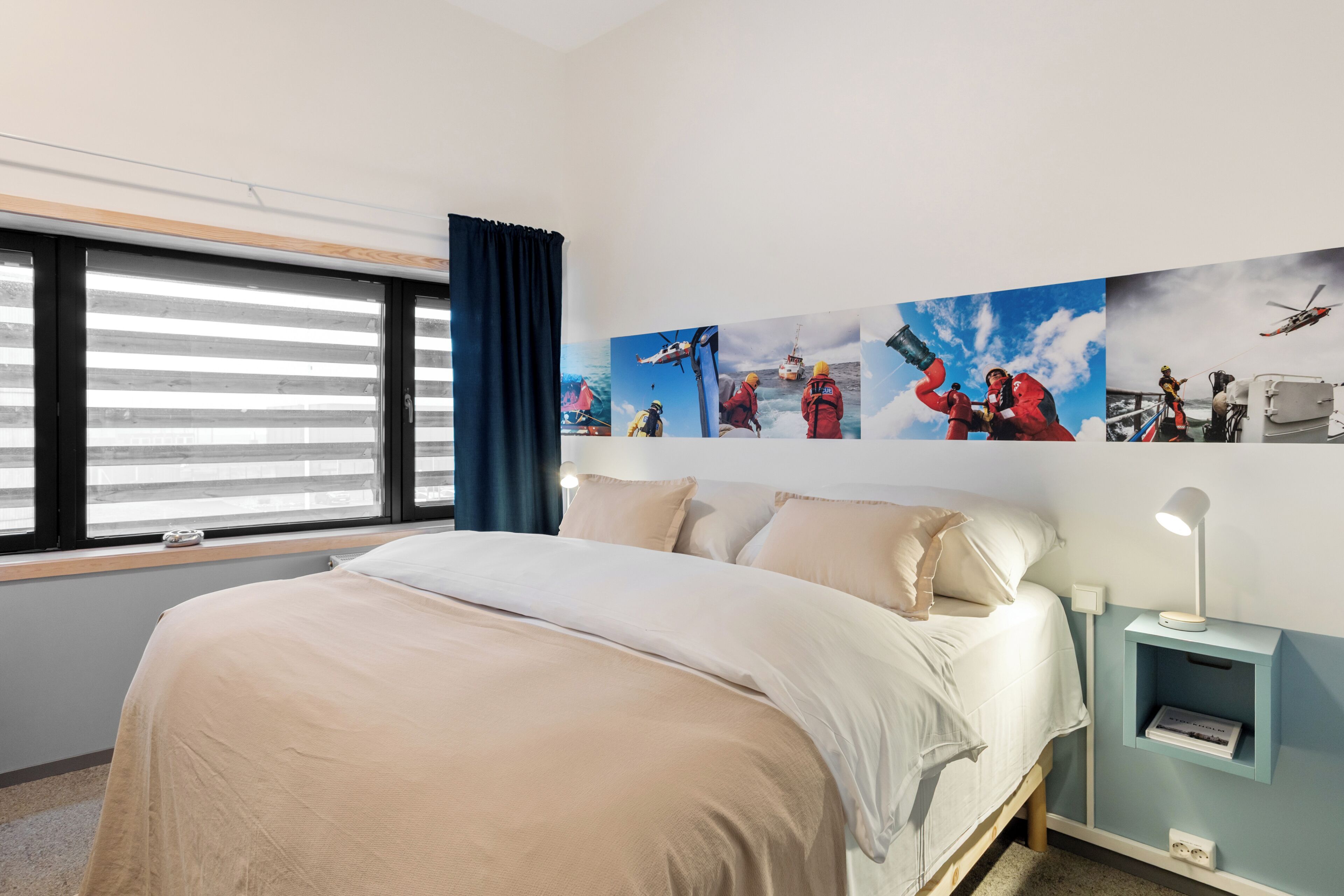 A bedroom with a bed, a window with blinds, a wall-mounted shelf with a lamp, and a series of photos depicting outdoor activities.