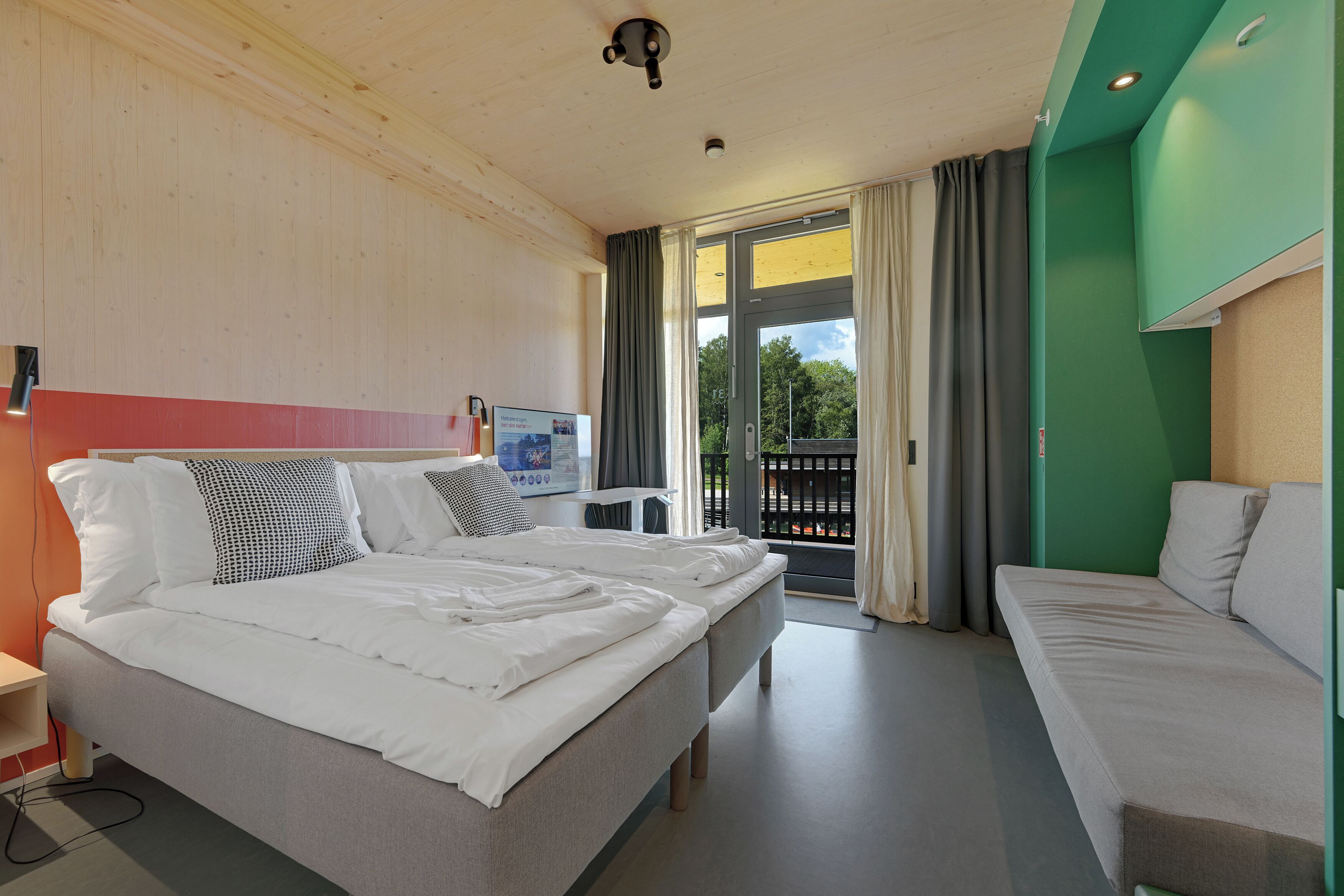 A hotel room with a bed, a sofa, a window with curtains, a wall-mounted TV, and a balcony.