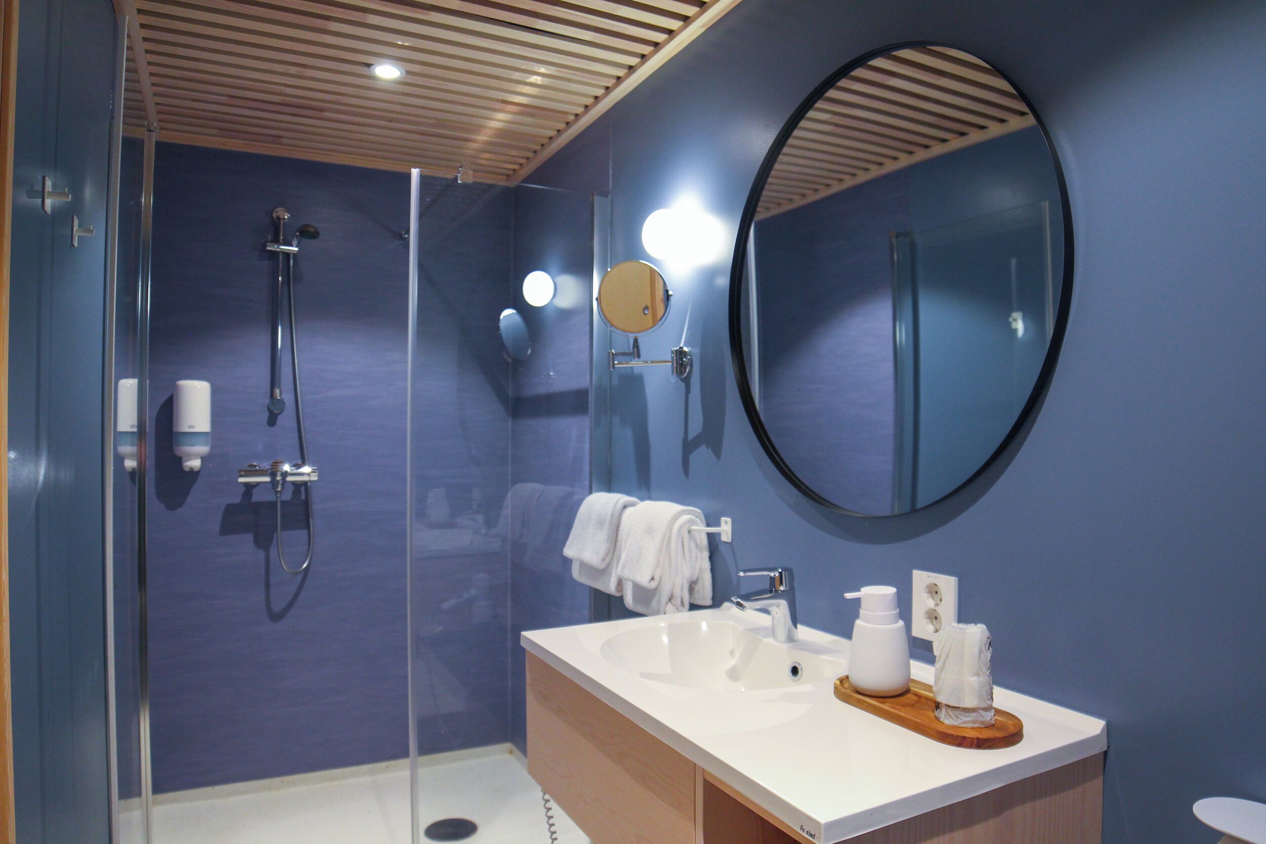 A modern bathroom with a glass-enclosed shower, a round mirror, and a white sink with a wooden tray.