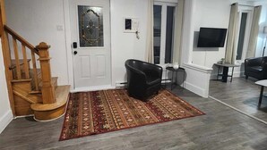 A spacious living room with a wooden staircase, a black leather sofa, a patterned rug, and a flat-screen TV mounted on the wall.