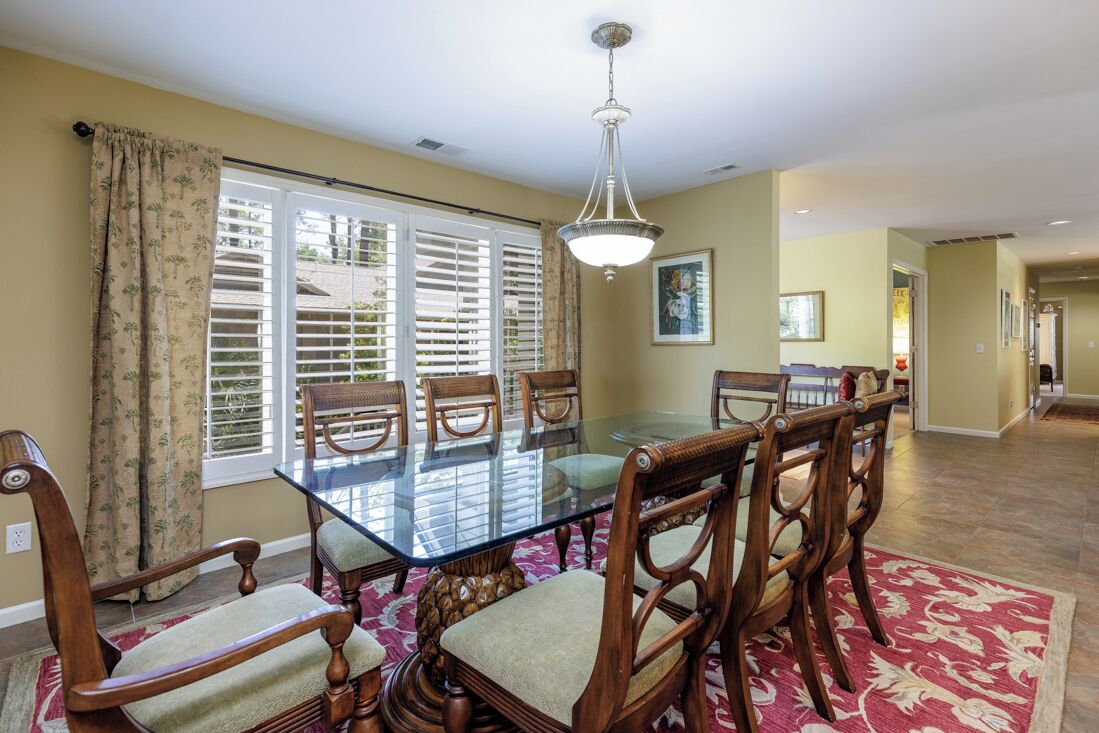 A dining area with a glass table and wooden chairs, a chandelier, and a view through a window with blinds.