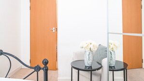 A bedroom with a bed, a bedside table with a vase, a wooden door, and a coat rack. - The Grand Serenity Suite (London)
