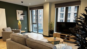 A modern living room with a grey sofa, a coffee table, and a TV. There is a large window with a view of buildings outside. - Spacious & Luxurious 2-bed Apartment in London (London)