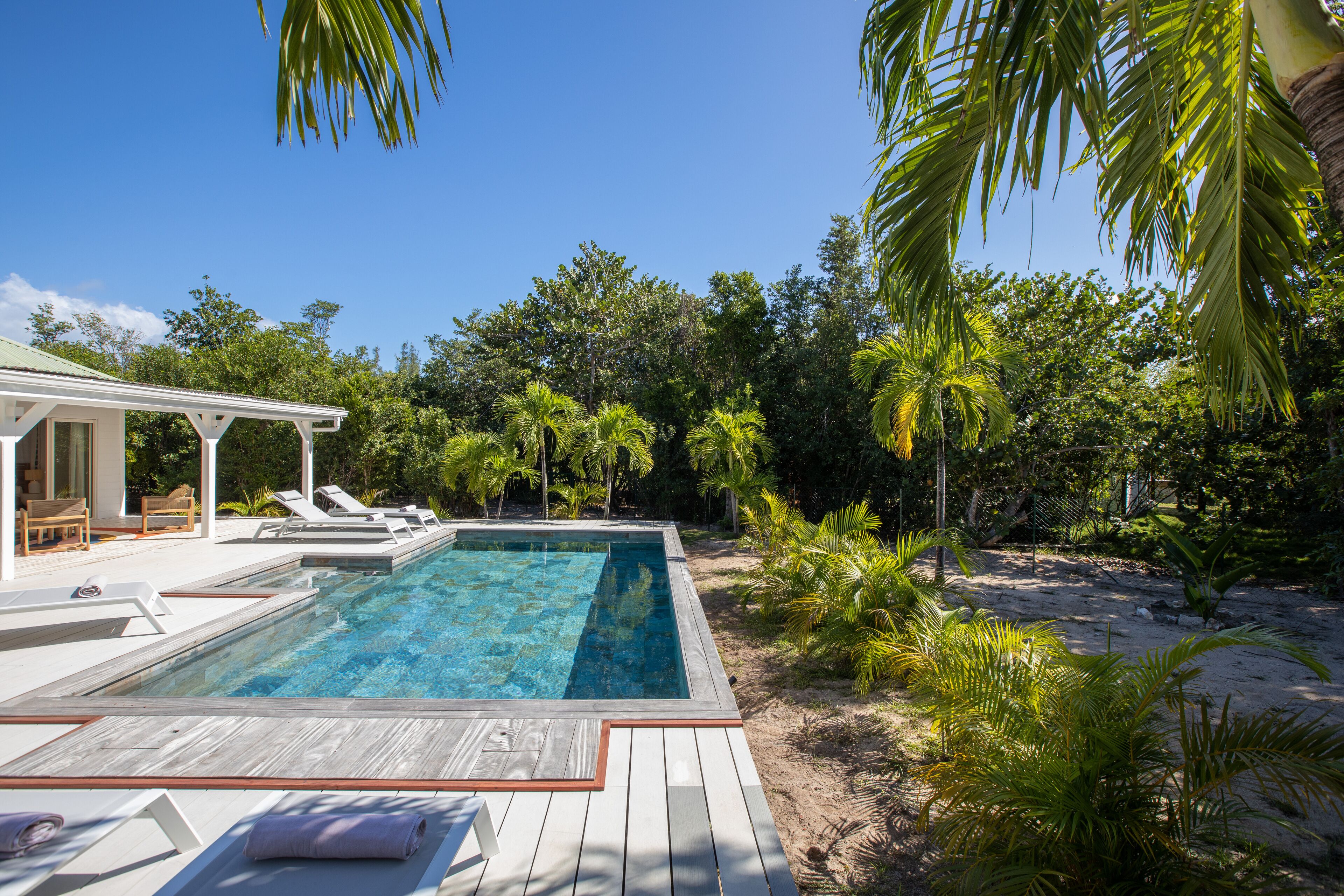 Une piscine dotée de chaises longues et d’un espace de repos couvert, le tout entouré d’une végétation tropicale et d’une plage de sable.