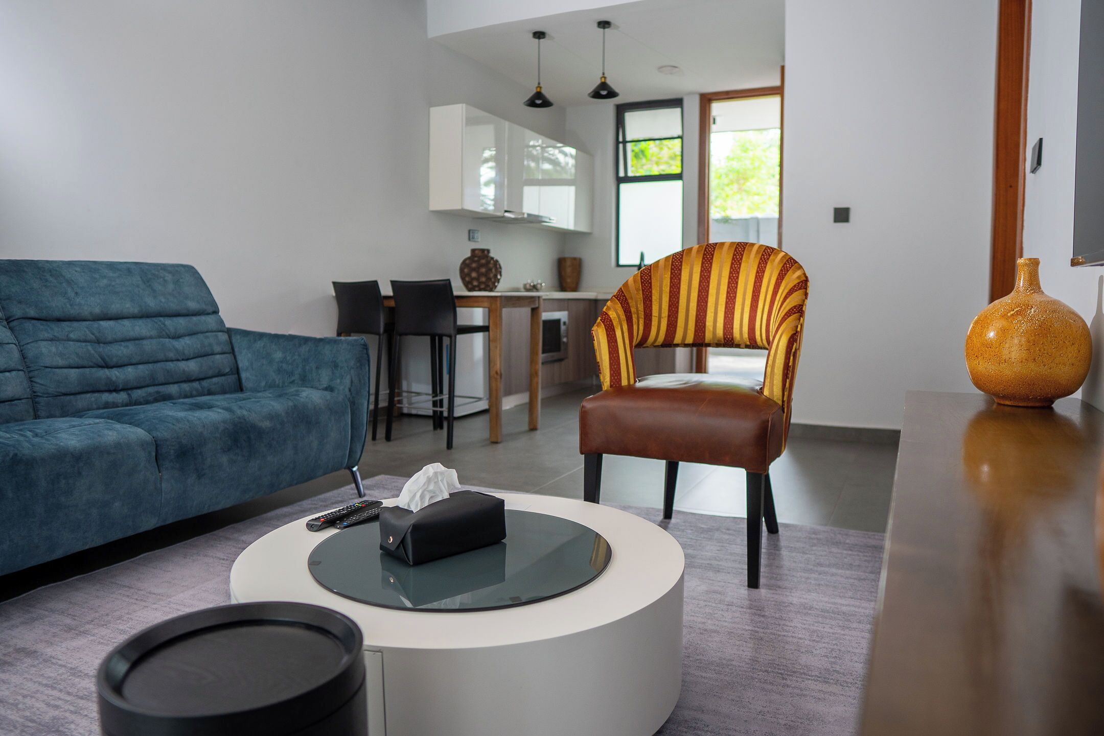 Una sala moderna con un sofá azul, una mesa de centro redonda, un sillón a rayas y un comedor con una mesa de madera y sillas.