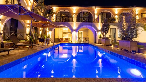 Un área junto a la piscina con tumbonas y un edificio con ventanas de arco y una iluminación cálida.