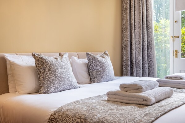 A hotel room with a large bed, patterned pillows, a neatly folded set of towels, and a view of greenery through the window.