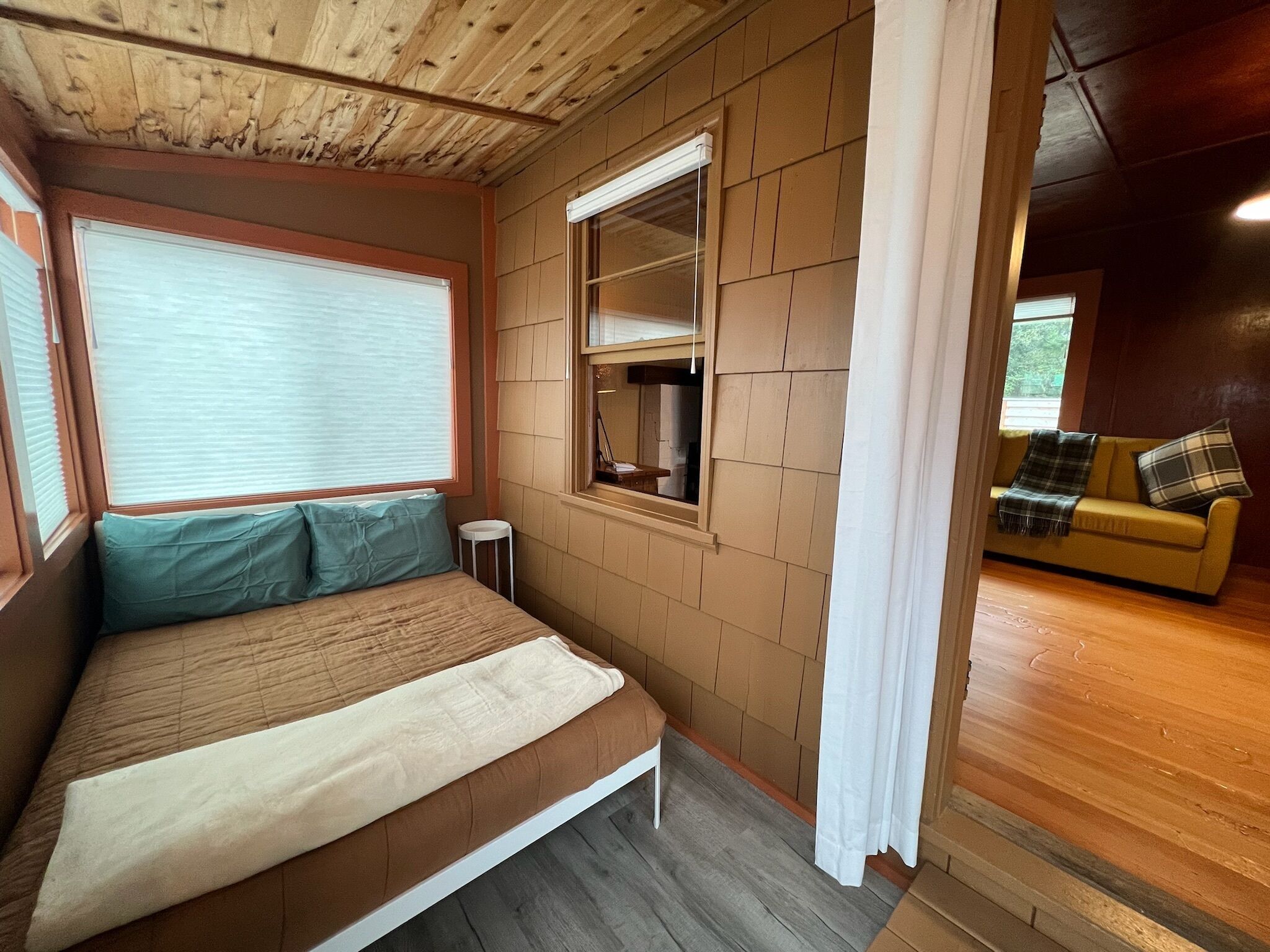 Una habitación con una cama, una mesita, un ventanal con persianas, un espejo y un sofá con una manta.