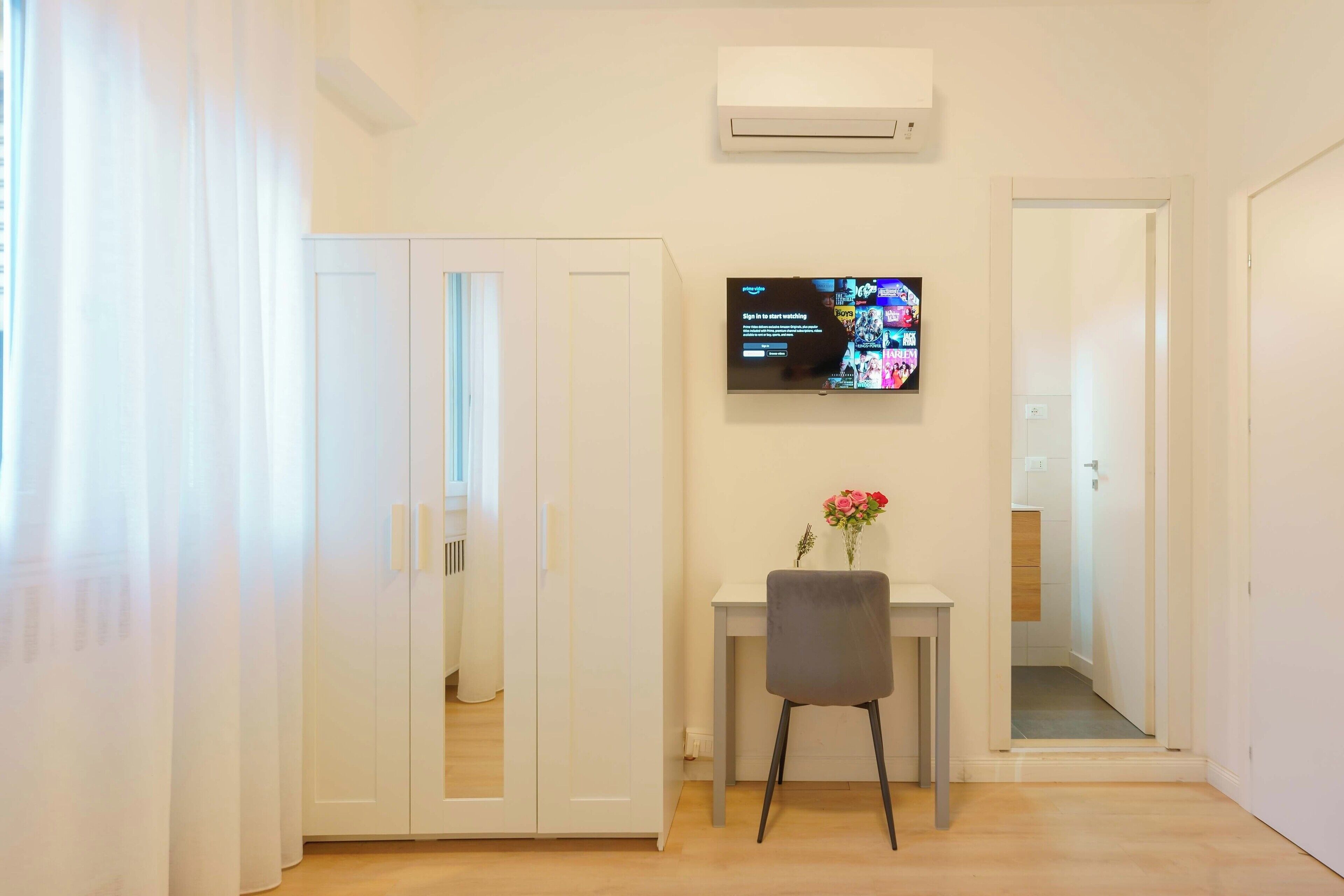 A small, bright room with a TV on the wall, a desk with a chair, and a wardrobe.