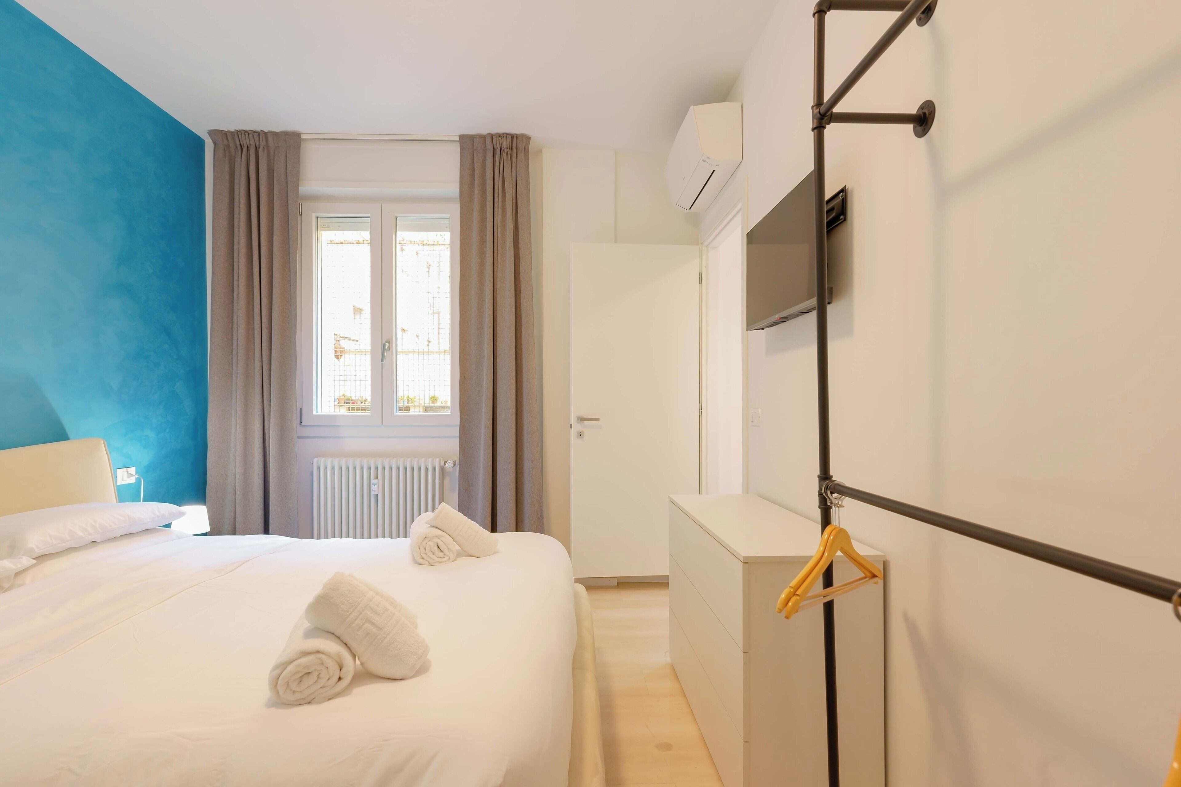 Une chambre d’hôtel avec un lit, une commode, un téléviseur fixé au mur et une fenêtre avec des rideaux.