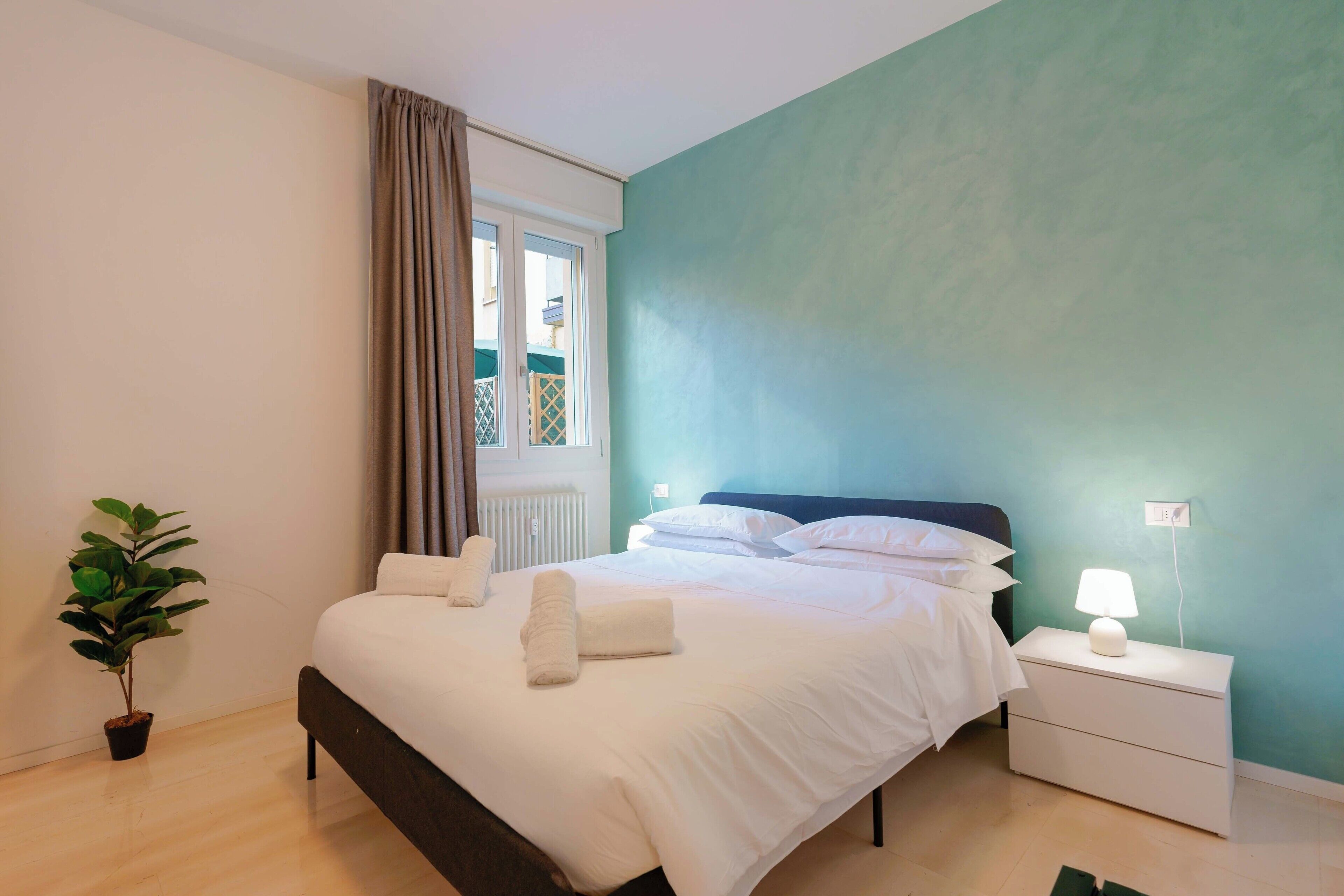 Una cama bien hecha con sábanas blancas y dos toallas enrolladas, una lámpara de noche, una planta en maceta y una ventana con cortinas.