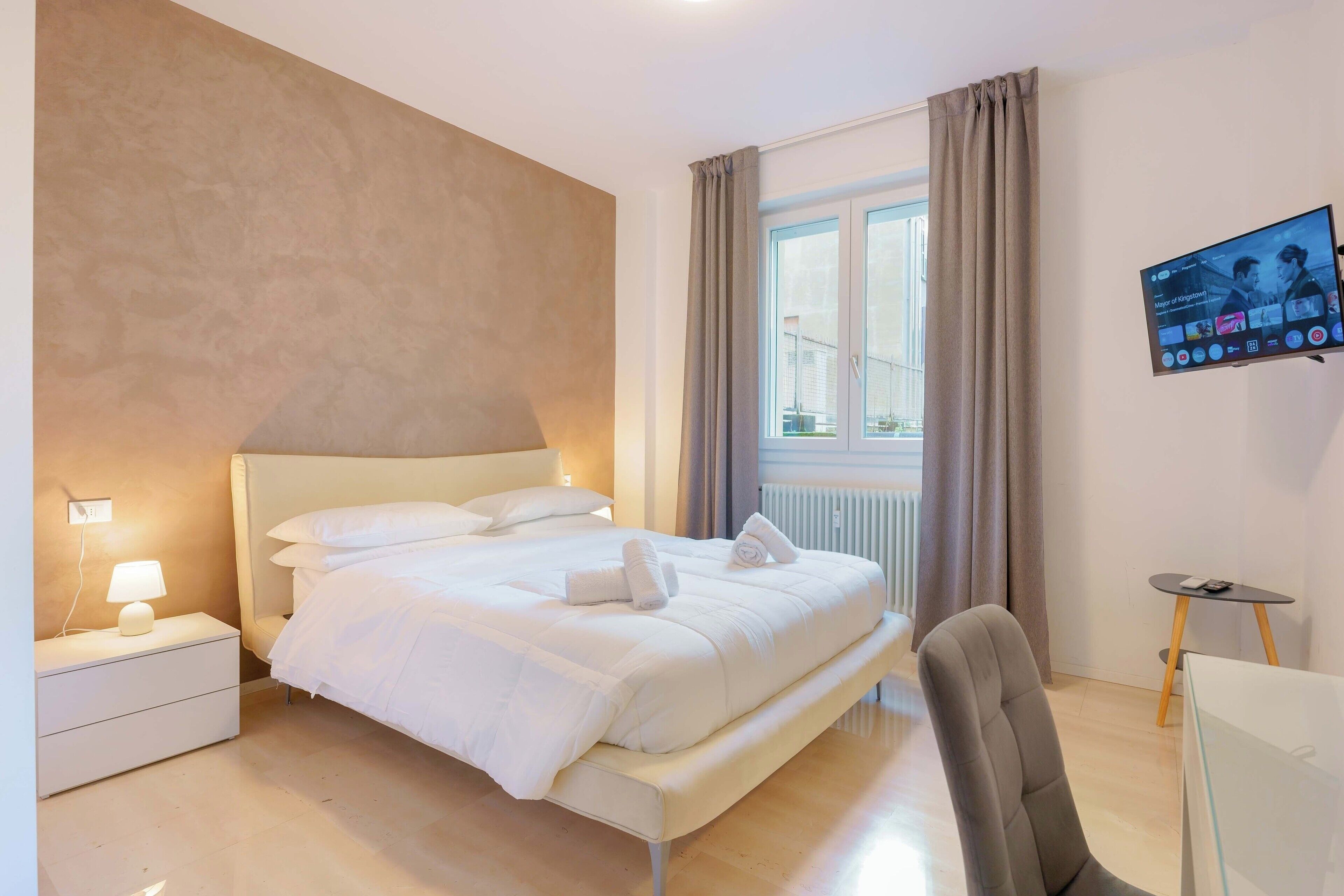 A modern hotel room with a large bed, a bedside table with a lamp, a TV mounted on the wall, and a view of the outdoors through a window.