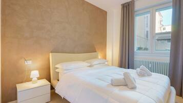 A neatly made bed with white linens and two rolled towels, a bedside lamp, and a window with curtains.