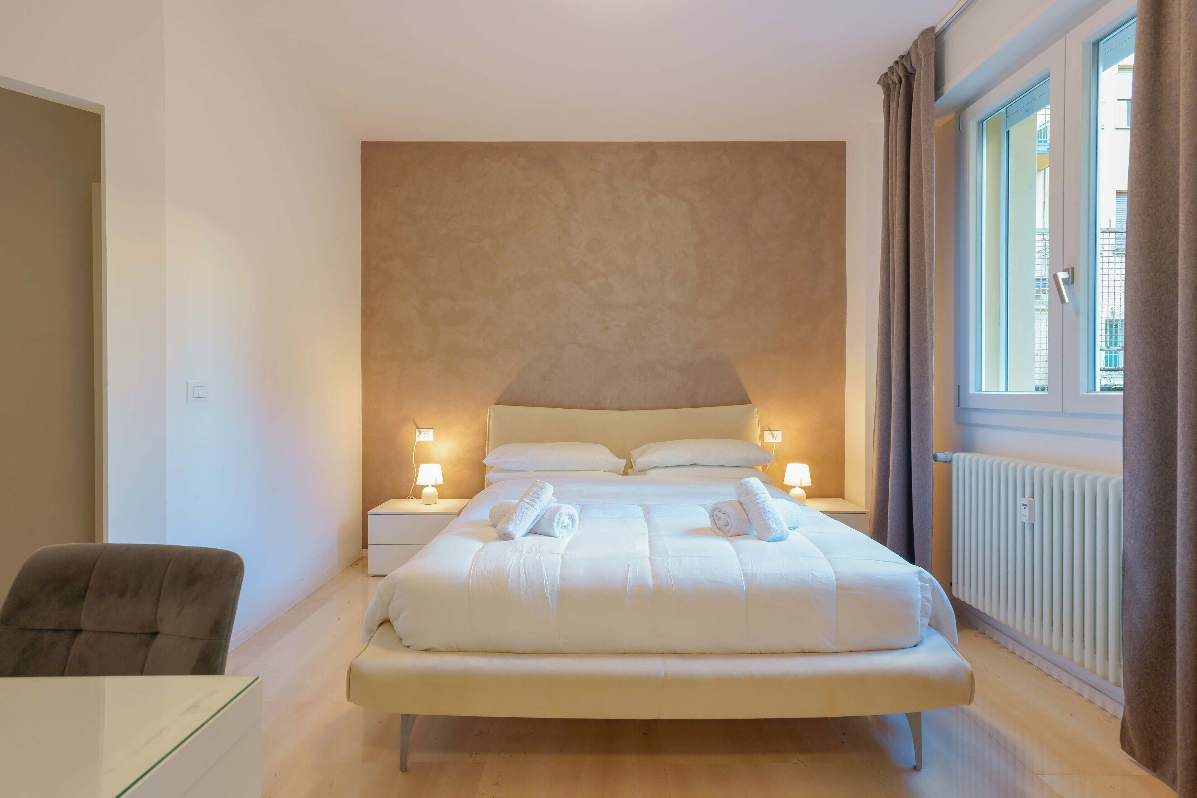 A bedroom with a large bed, bedside lamps, a chair, and a window with curtains.