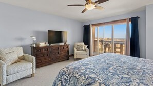 A bedroom with a bed, a dresser, a TV, two chairs, a ceiling fan, and a large window with curtains. - Sea Mist: Oceanfront | Pool and Tennis Access (Nags Head)