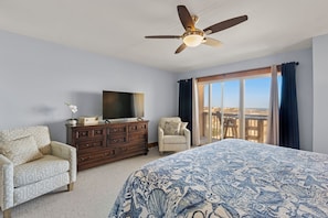 A bedroom with a bed, a dresser, a TV, two chairs, a ceiling fan, and a large window with curtains.